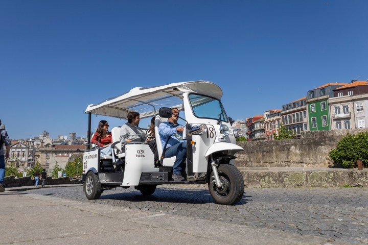 Open-air white tuk-tuk with four passengers rides on cobblestone street in sunny urban area.
