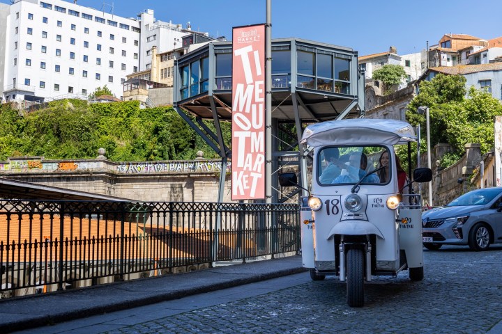 White tuk-tuk on cobblestone street near a market sign in an urban area.