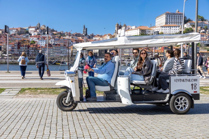 Group of people on a white tuk-tuk by a riverside, cityscape in background.