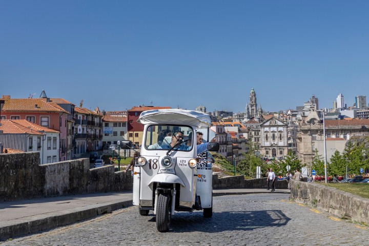 White tuk-tuk on cobblestone street with cityscape and blue sky in background.