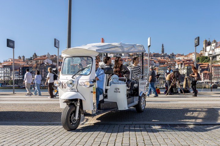 Group in white tuk-tuk on a sunny day with cityscape in background.