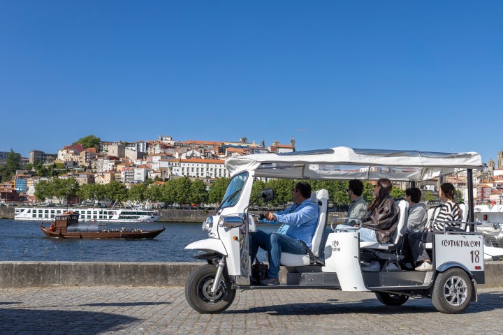 Tourists in a white tuk-tuk by a riverside with a cityscape view.