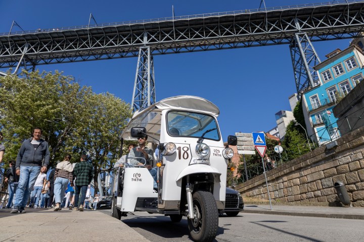 White tuk-tuk on city street with bridge overhead on a sunny day.