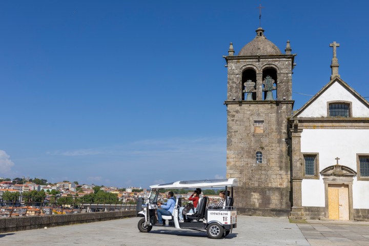 Tourists in a white tuk-tuk near an old bell tower on a sunny day.