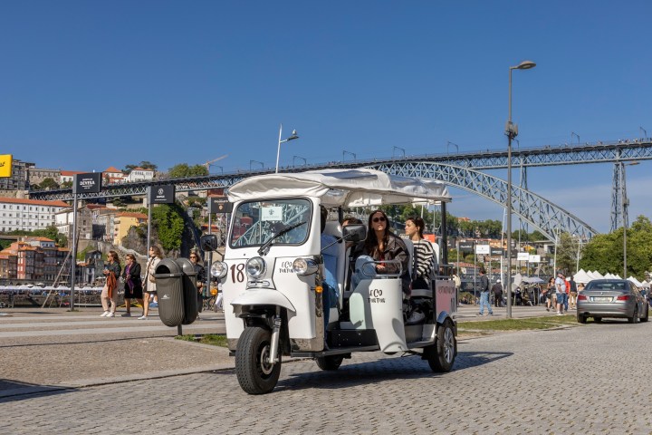 White tuk-tuk with passengers in front of a bridge on a sunny day.