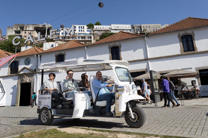 A white tuk-tuk with passengers on a cobblestone street near buildings and a CALÉM sign.