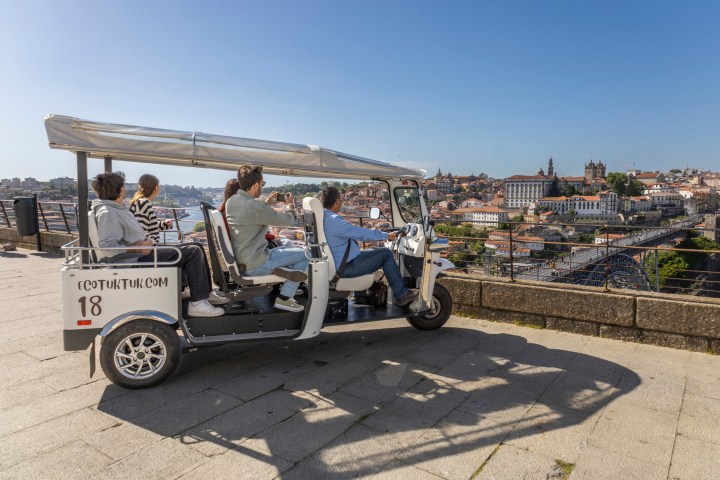 Tourists in a white tuk-tuk overlooking a city skyline and river.